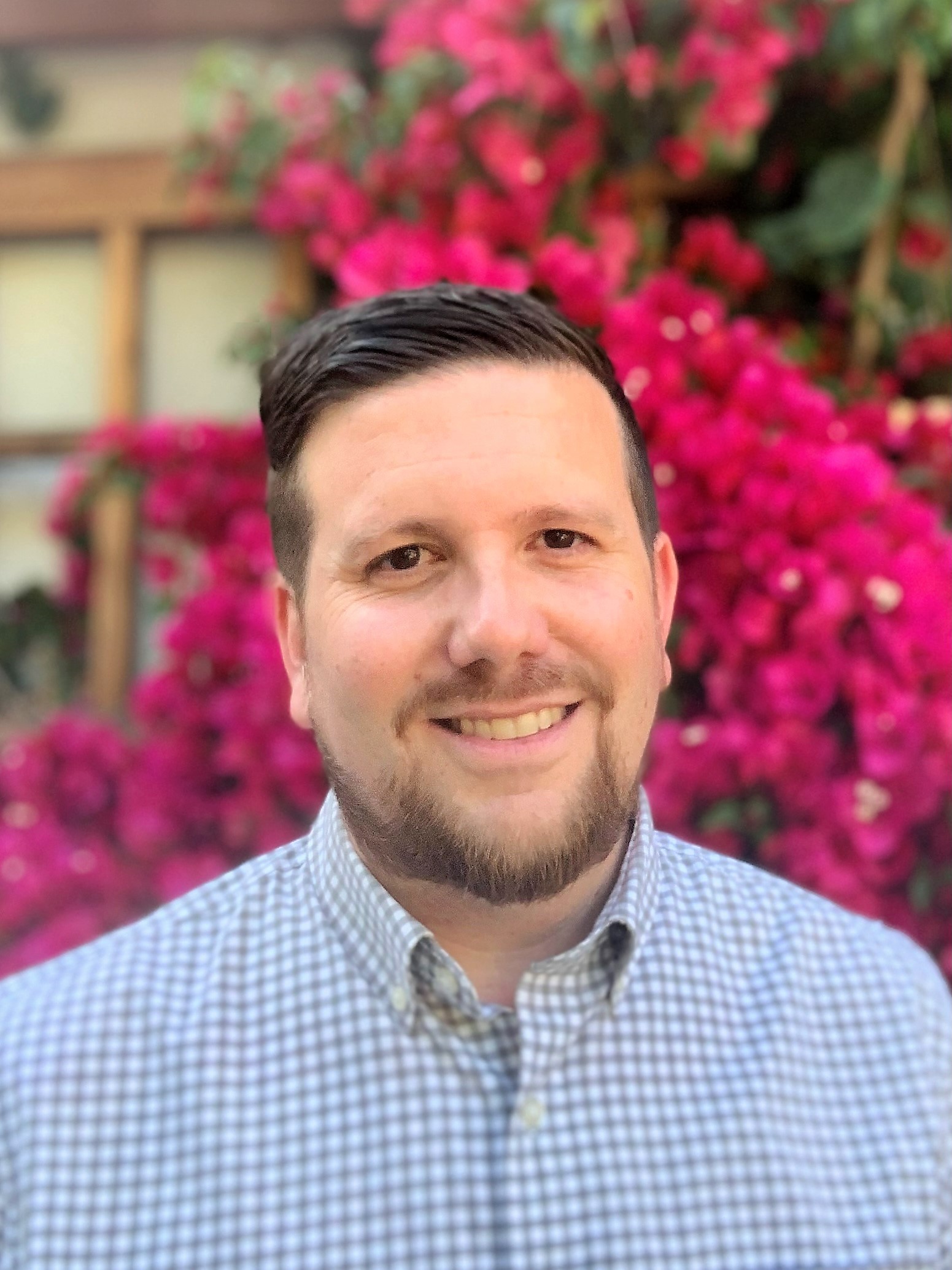 photo of prof Daniel Zazueta a man with brown hair and a beard wearing a blue checkered shirt with a collar he is standing in front of a flowering pink bougainvillea plant