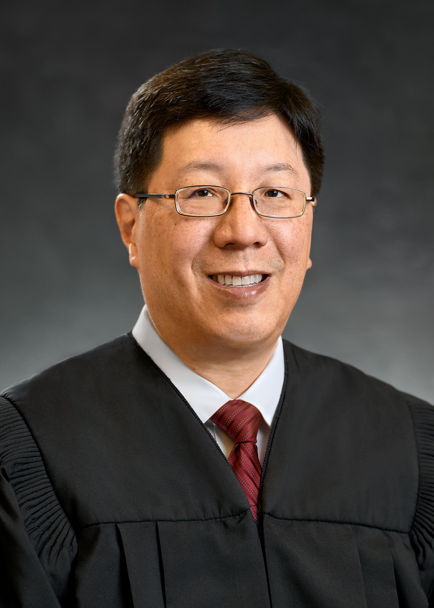 headshot of justice danny chou an asian man with black hair and classes wearing a black judicial robe white button down shirt and a maroon tie