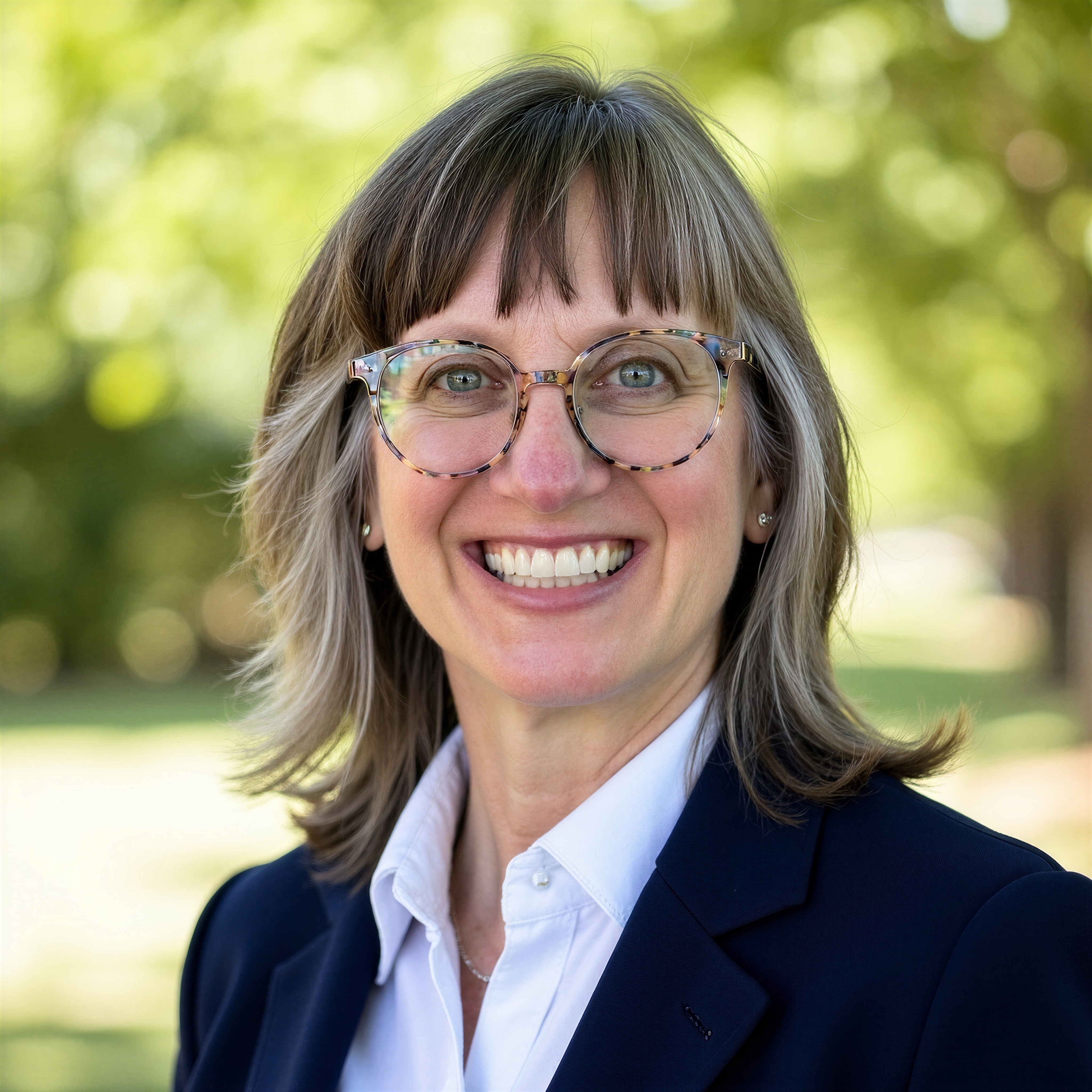 photo of prof laura norris a smiling white woman with brown and grey hair with bangs, she is wearing colorful glasses, a white collared button down shirt and a blue blazer the background is greenery