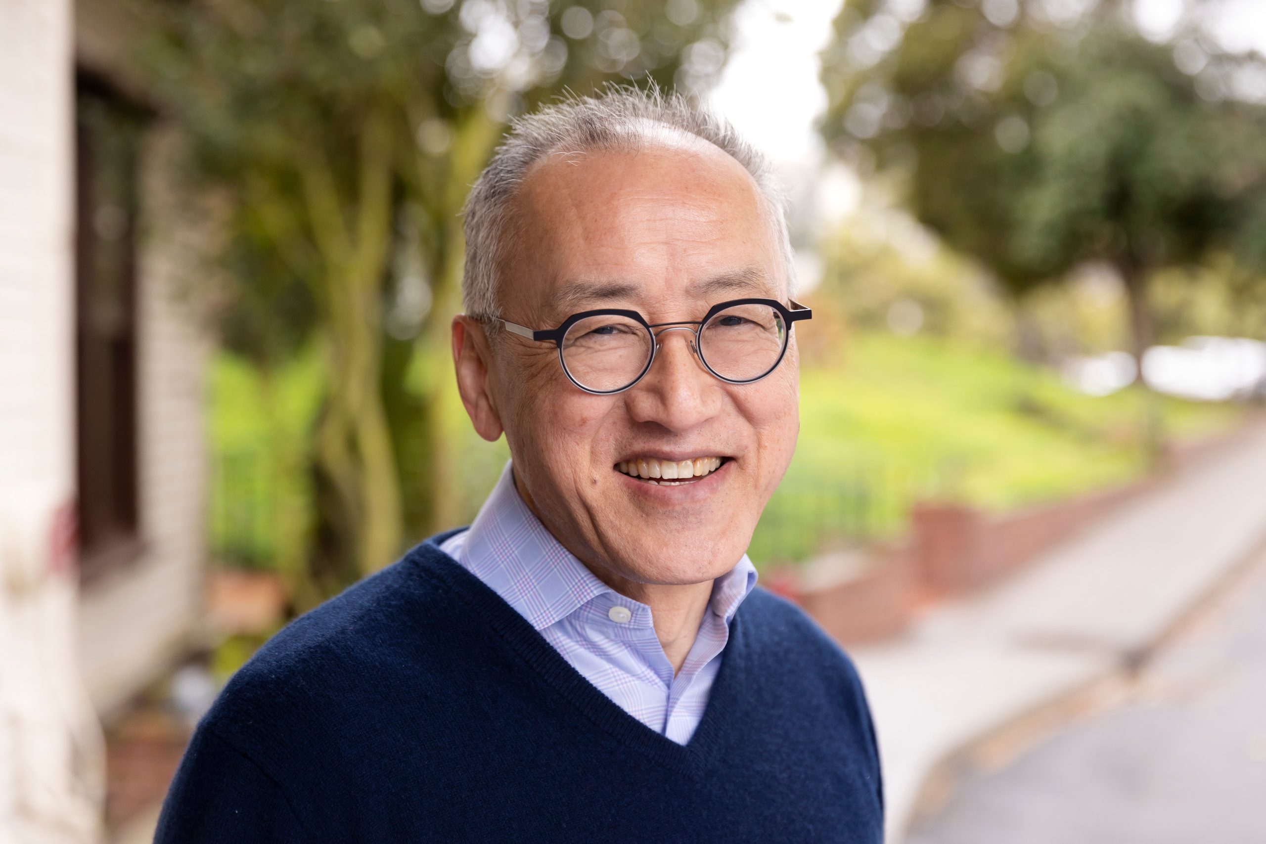 a photo of prof larry low, an asian man wearing glasses a blue collared shirt and blue sweater on a backdrop of plants