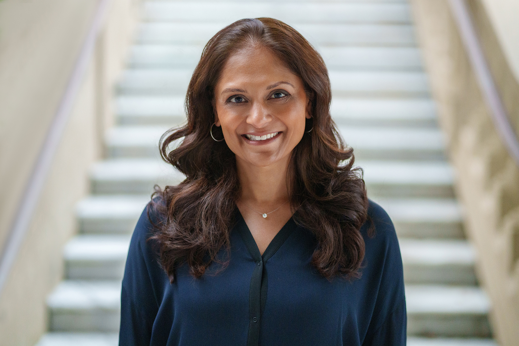 photo of prof sejal patel a smiling woman with redish brown hair wearing a navy shirt and a small necklace the background is a set of white stairs