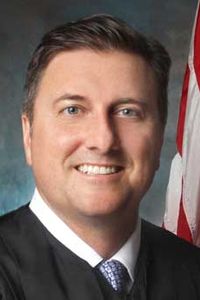 headshot of prof wes porter a white man with brown hair wearing a white button down shirt, blue tie and judicial robes with a small corner of the stripes from the American flag in the background