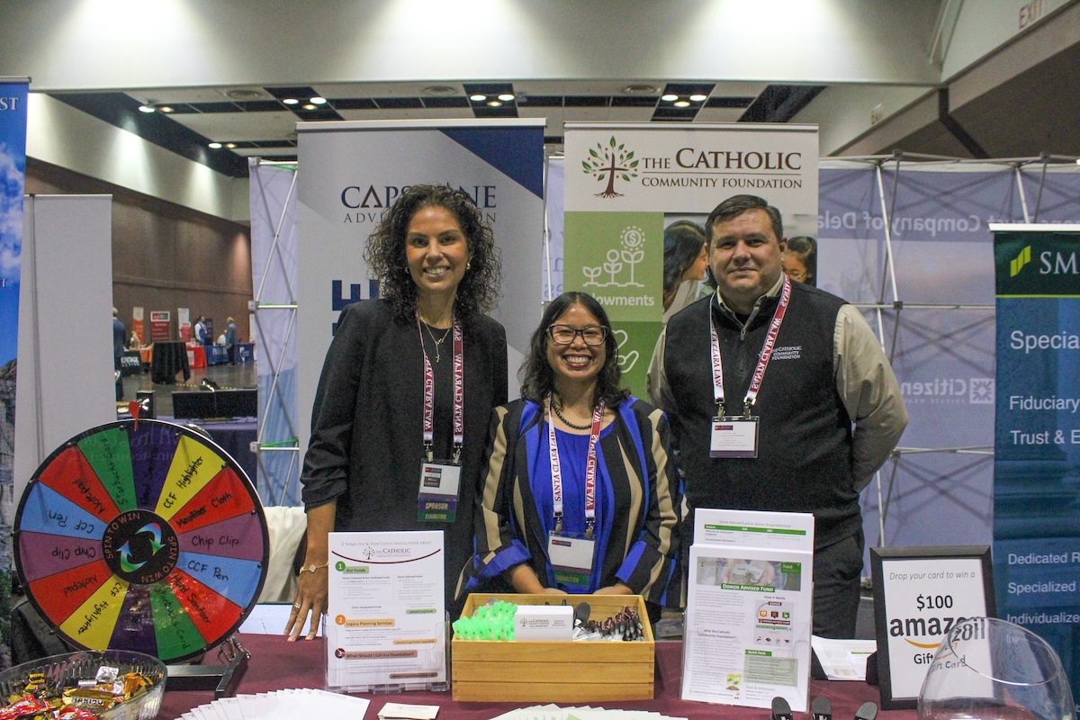 2 women and a man standing in front of a Catholic Charities booth