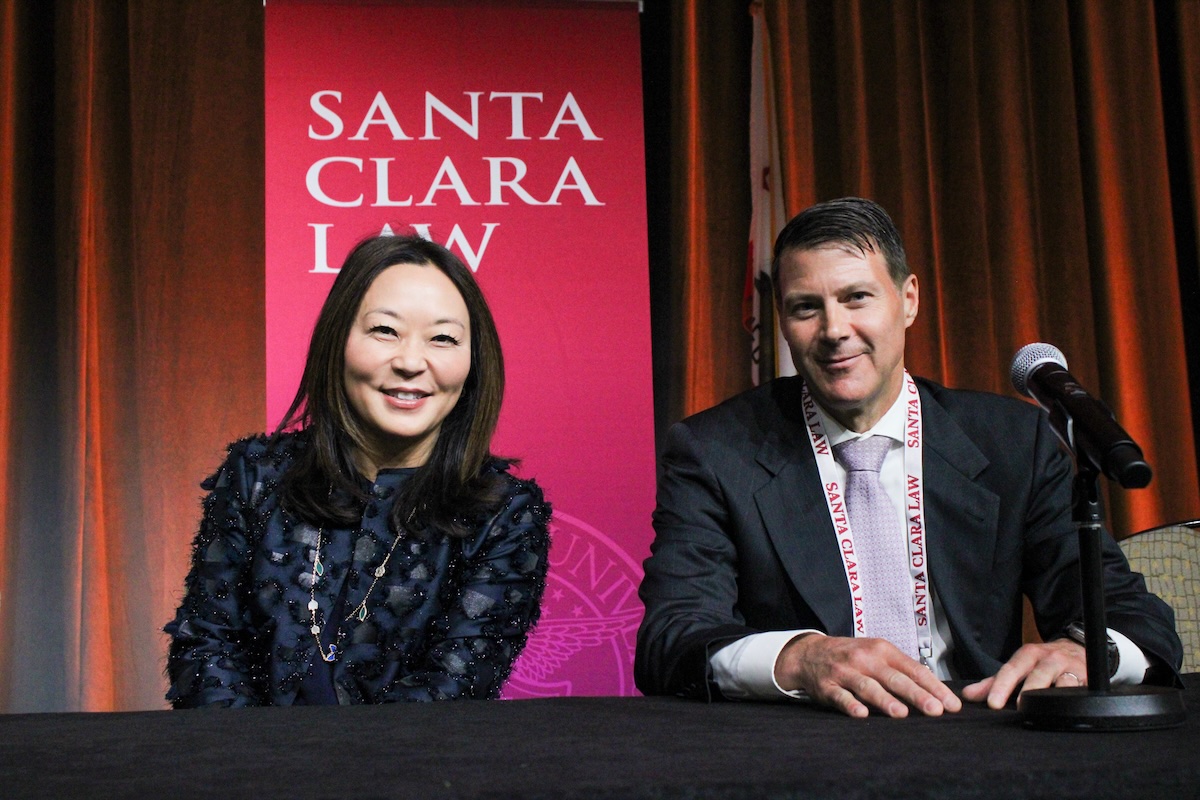 a man and woman smiling in front of a red Santa Clara law banner