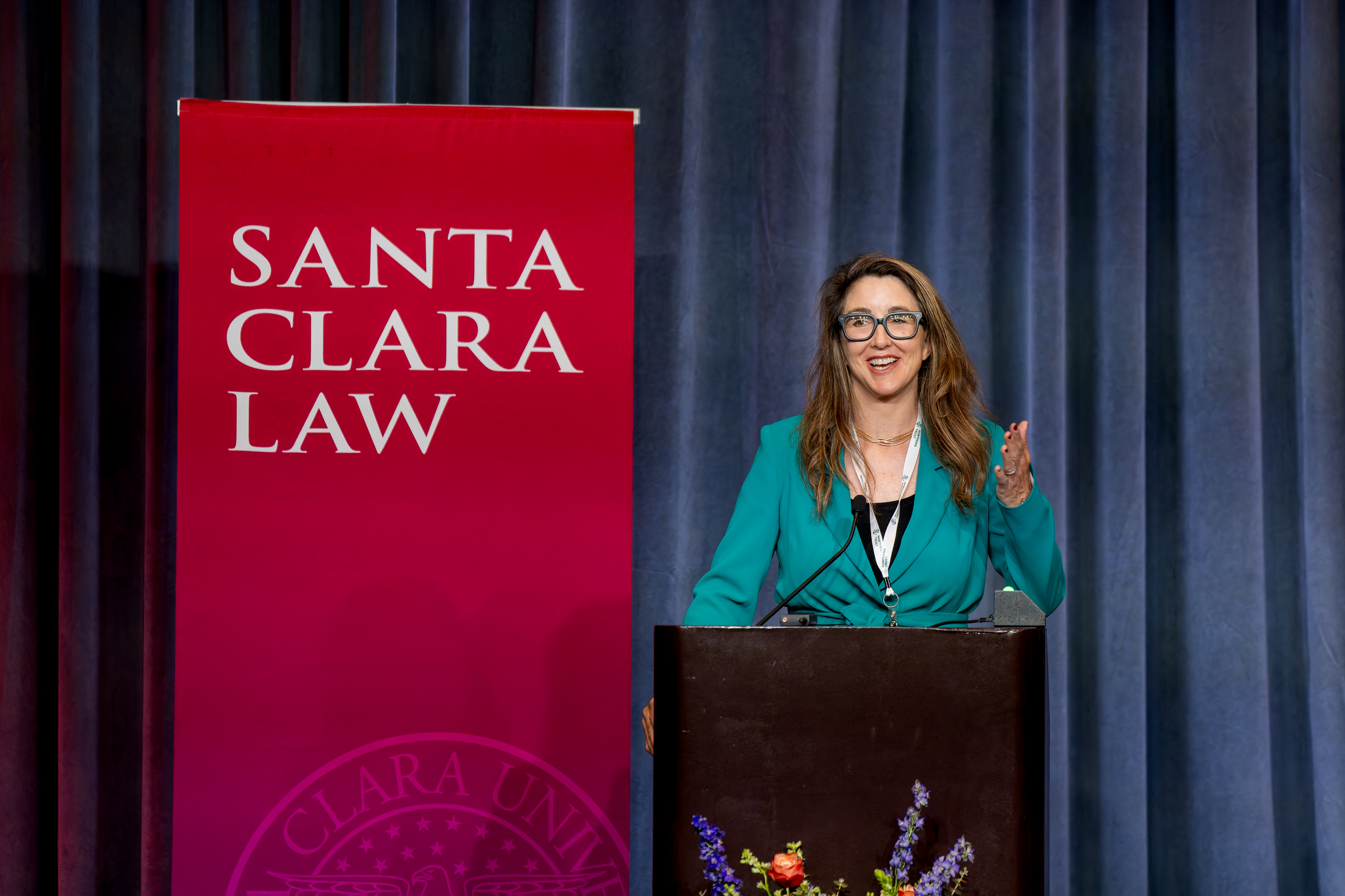 A lady in a greenish blue suit at a front of the stage talking at an event.