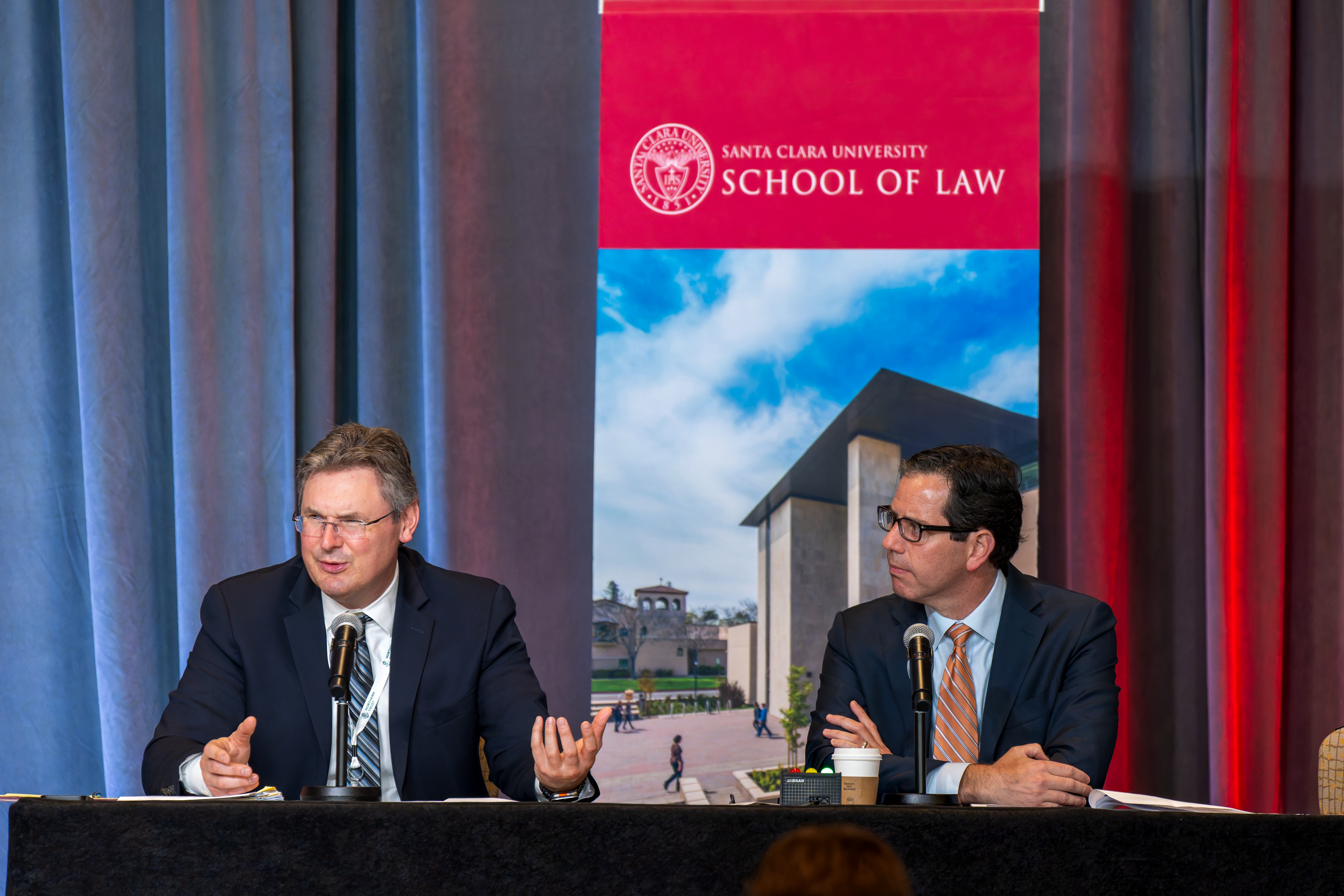 two men at a table on the front stage at an event speaking to a crowd.