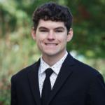 photo of law student standing in garden wearing a black suit, with a black tie and a white and black polka dot shirt, he has curly brown hair
