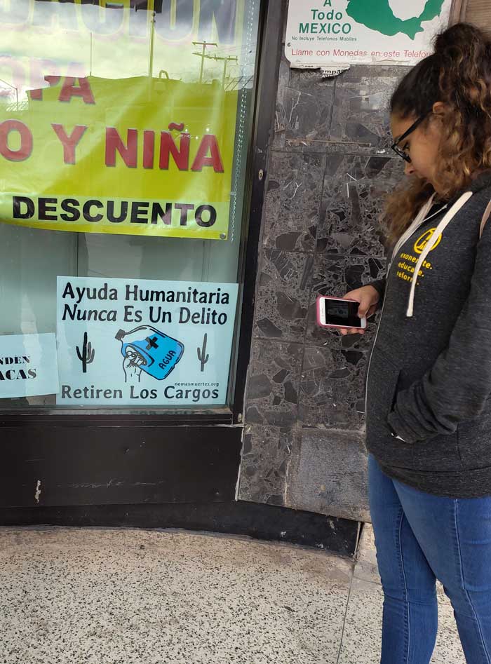 Ruby Palomares reading a sign in the window of a store.  It says “Humanitarian Aid Is Never a Crime.  Withdraw the Charges.” And it shows a bottle of water.